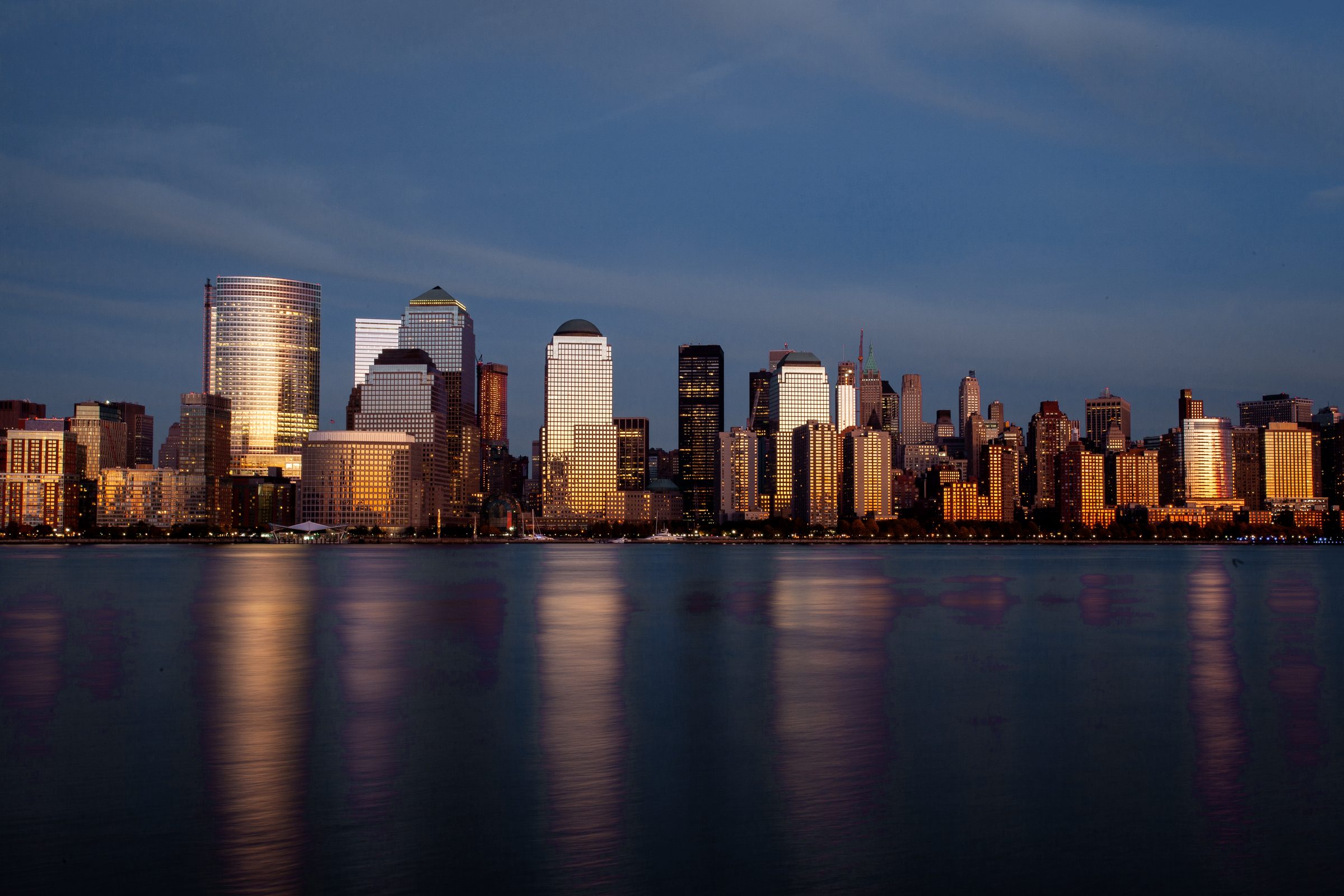 A downtown skyline of glass towers at dusk, lit windows reflected in calm water