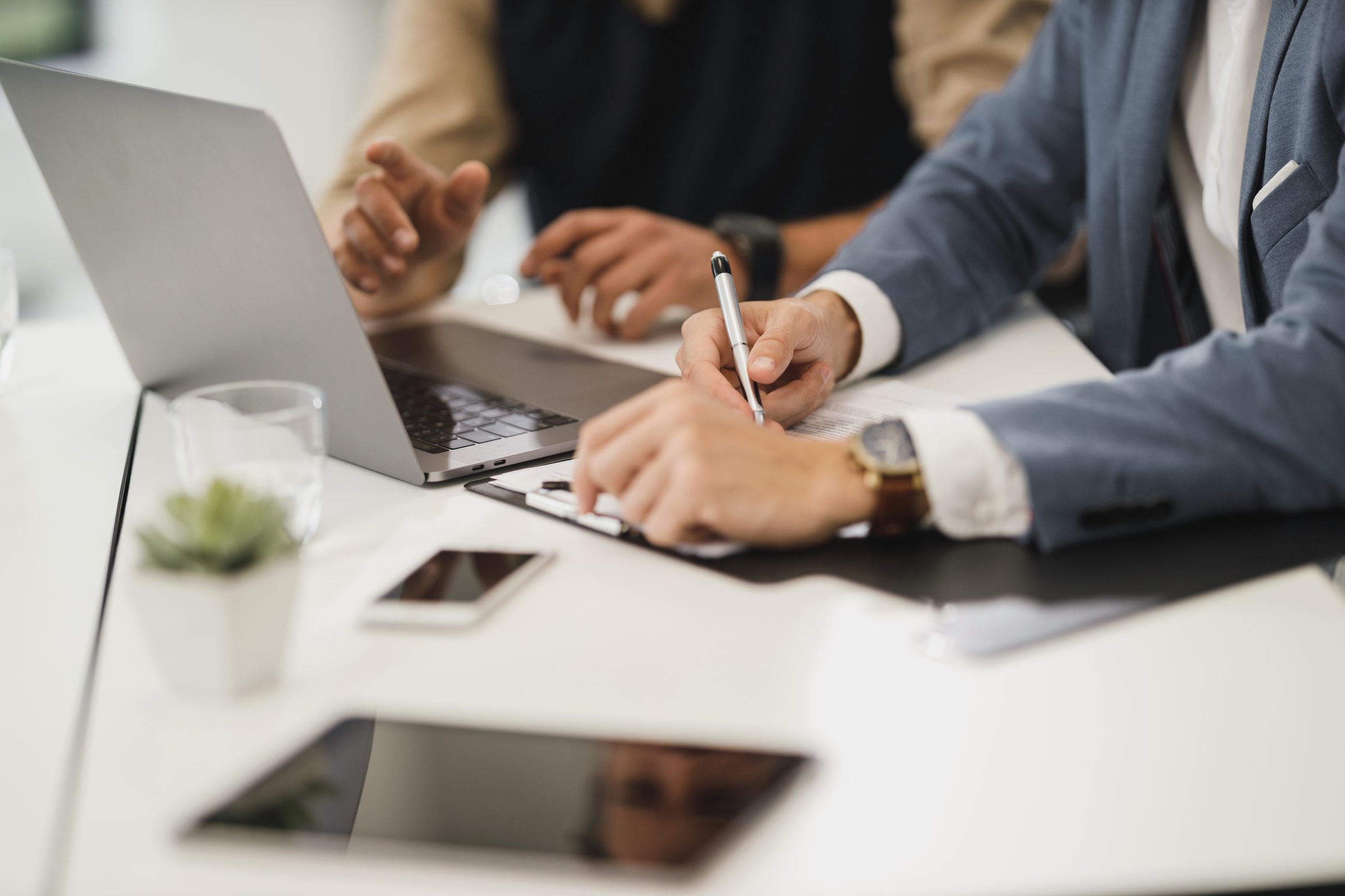 Two professionals at a bright office table reviewing documents beside a laptop, pen in hand