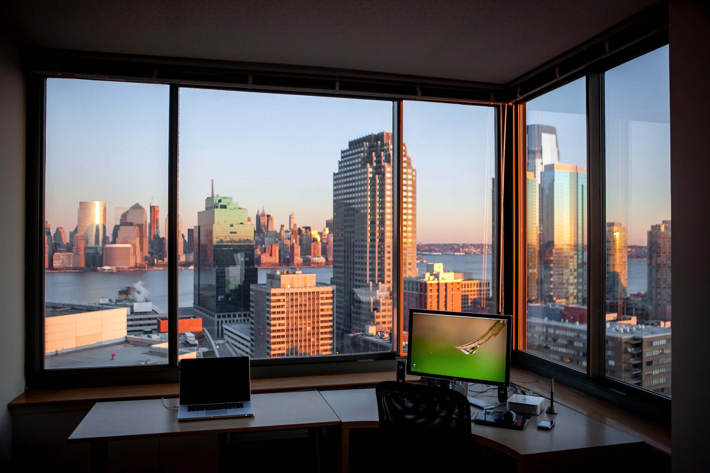 A corner office with a large display and floor-to-ceiling windows onto a city skyline at dusk